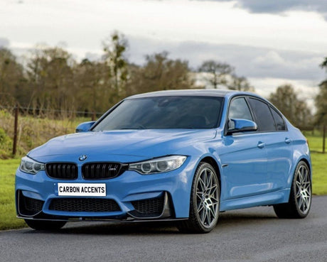 A blue sports sedan is parked on a road, with trees and clouds in the background. The license plate reads "CARBON ACCENTS."