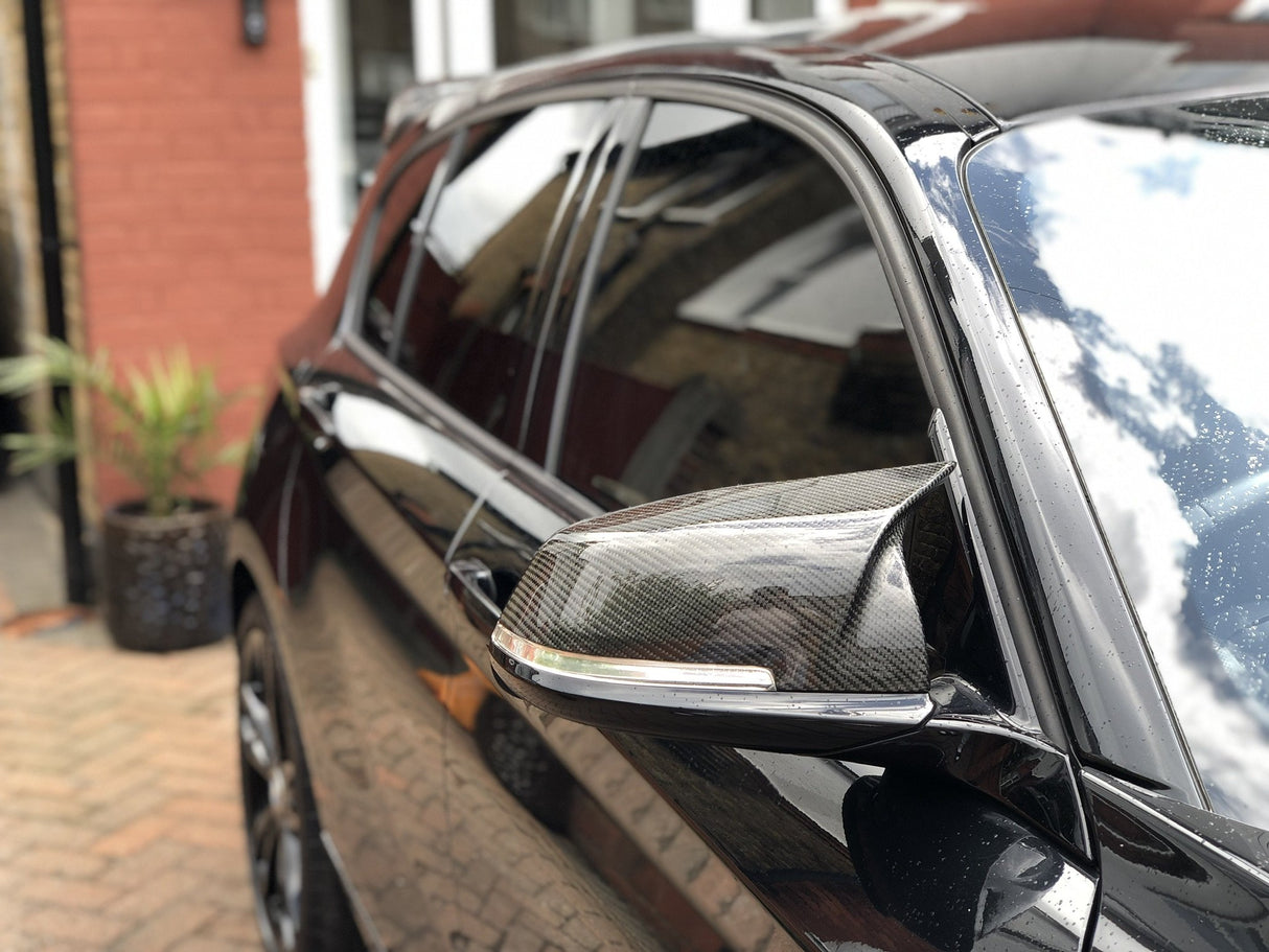 A shiny black car's side mirror reflects the sky, parked beside a red-brick building with a plant pot on a paved driveway.