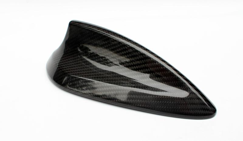 A sleek, black carbon-fiber car antenna rests on a white surface, showcasing a streamlined, shark-fin design that reflects light, highlighting its glossy texture and modern aesthetics.