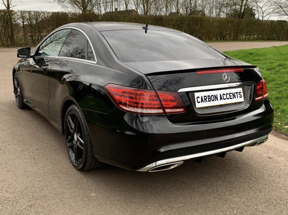 A black luxury car is parked on a road, with "CARBON ACCENTS" on the license plate. The car is set against a backdrop of grass and trees.