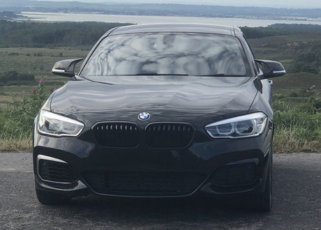 A black BMW car is parked on a road, facing forward, with a hilly landscape and a large body of water in the background.