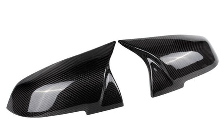 Two sleek, carbon fiber side mirror covers are angled toward each other, showcasing a reflective surface and intricate weave pattern against a plain white background.