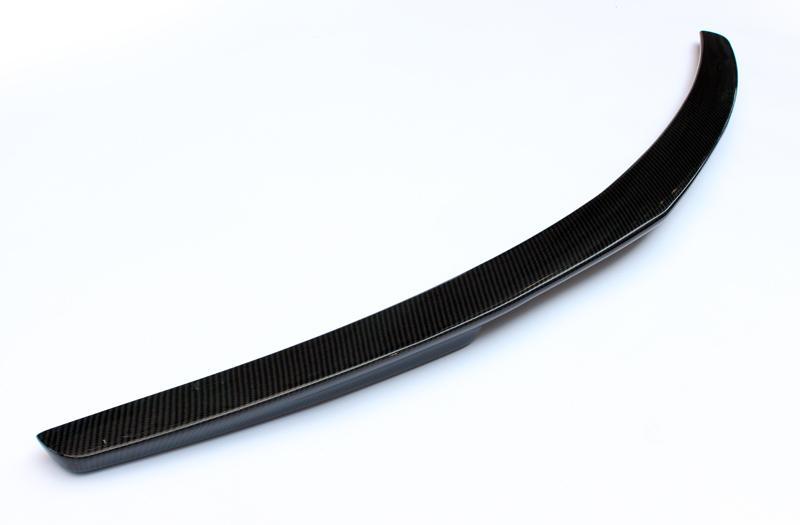 A curved, black carbon fiber car spoiler rests on a white surface, showcasing its sleek, aerodynamic design.