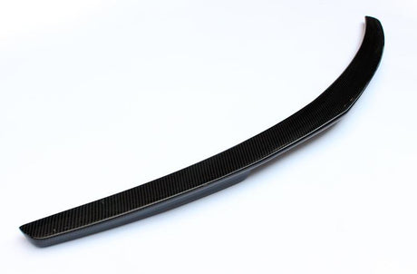 A curved, black carbon fiber car spoiler rests on a white surface, showcasing its sleek, aerodynamic design.