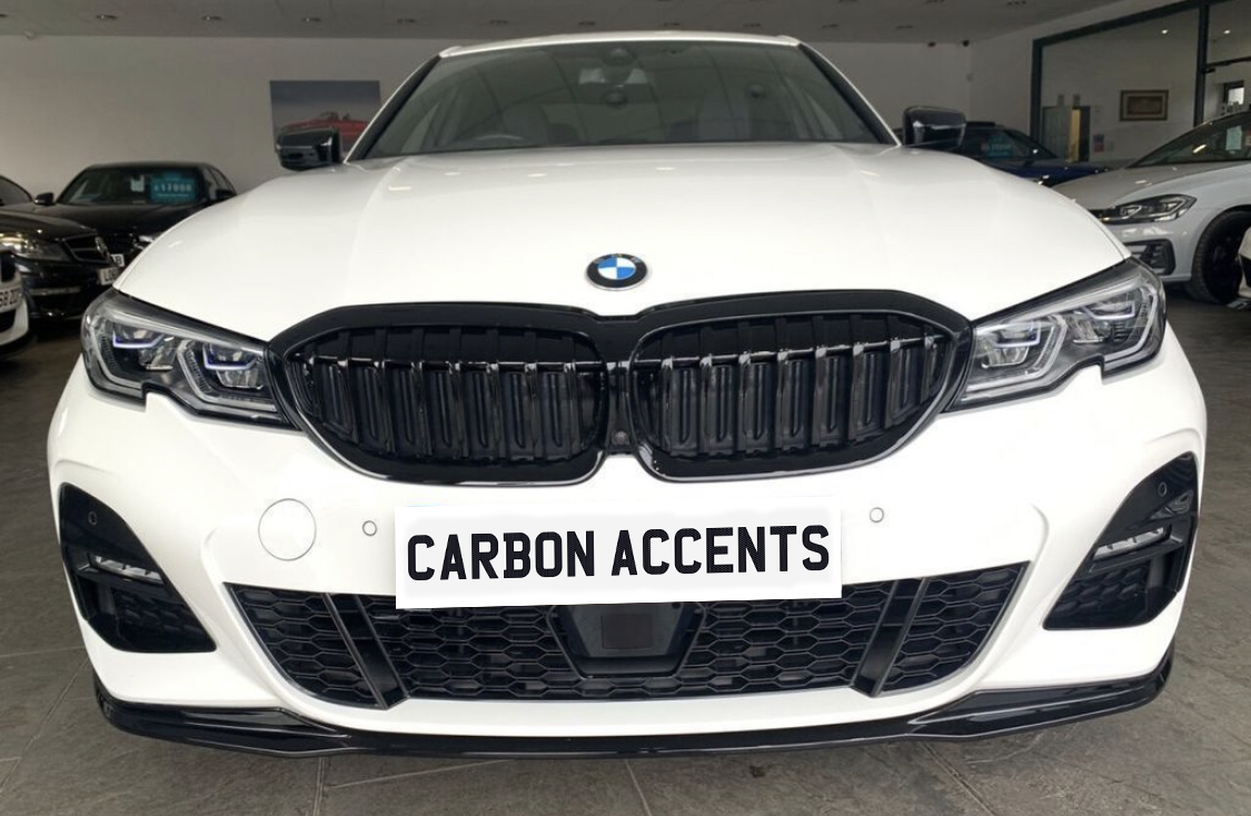 A white BMW car displays "CARBON ACCENTS" on the front license plate. The vehicle is parked indoors, surrounded by other cars, with visible headlights and kidney grille design.