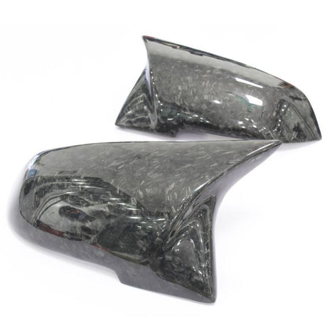 Two glossy, carbon fiber car side mirror covers with a pattern, resting on a white surface, reflecting light subtly across their surface.