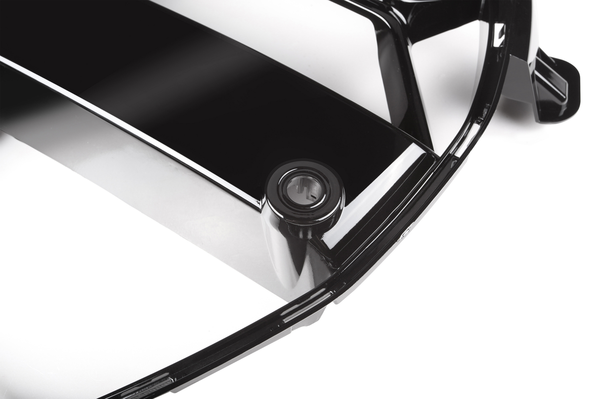 A glossy black plastic car part, possibly a grille, features a circular detail set within it. The surrounding area is a plain white background, highlighting the sheen of the part.