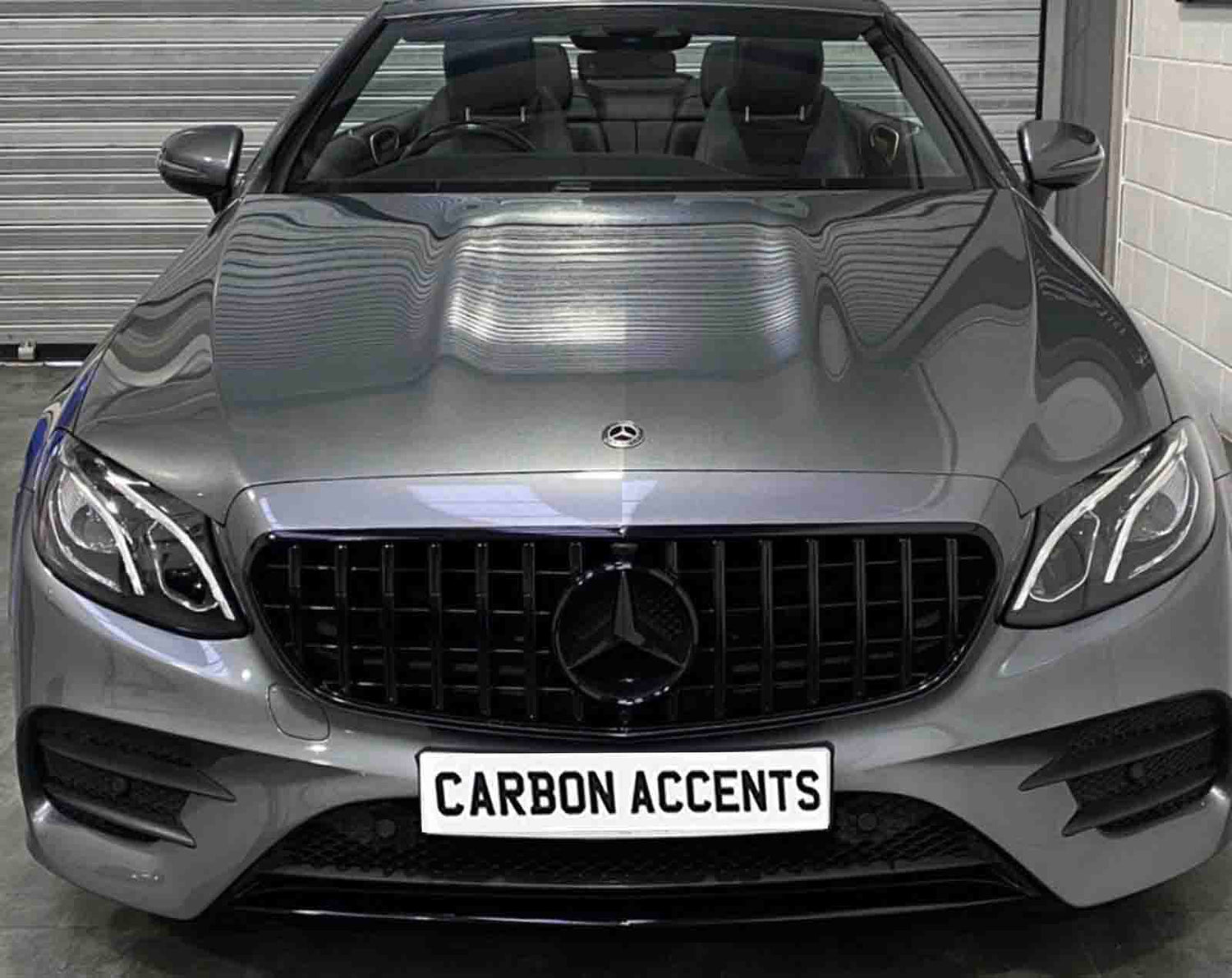 A sleek, gray car with a prominent grille and distinctive headlights faces forward in a garage. The license plate reads "CARBON ACCENTS," with surrounding concrete walls and a metal shutter backdrop.
