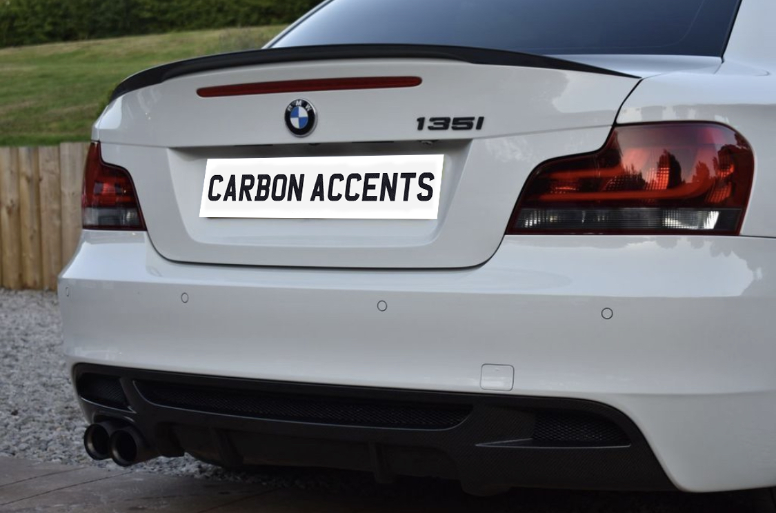 The car's rear features a white BMW 135i, with carbon fiber accents and "CARBON ACCENTS" license plate text. It is parked on gravel, with a wooden fence and grass in the background.