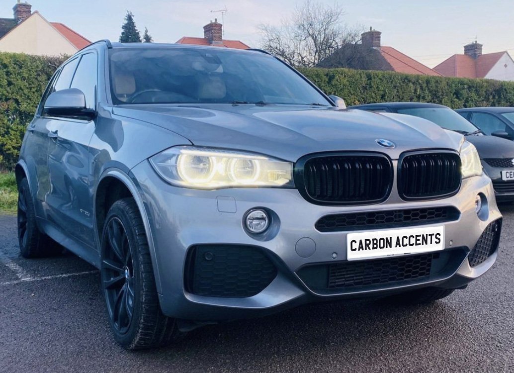 A silver SUV with illuminated headlights is parked on a paved surface. It features dark wheels and a front grilles. Surrounding are hedges and houses. Text on the plate reads “CARBON ACCENTS.”