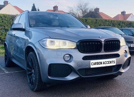 A silver SUV with illuminated headlights is parked on a paved surface. It features dark wheels and a front grilles. Surrounding are hedges and houses. Text on the plate reads “CARBON ACCENTS.”