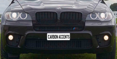 A black SUV's front view, with illuminated headlights and fog lights, is parked on grass. The license plate reads "CARBON ACCENTS."