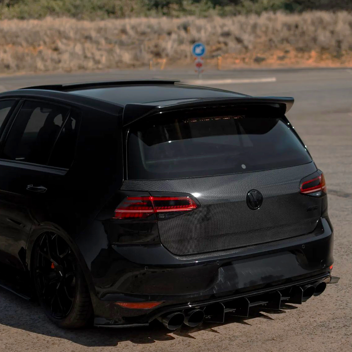 A black hatchback car is parked on a pavement with a carbon fiber rear, featuring prominent tailpipes and a roof spoiler, surrounded by a dry, grassy landscape.