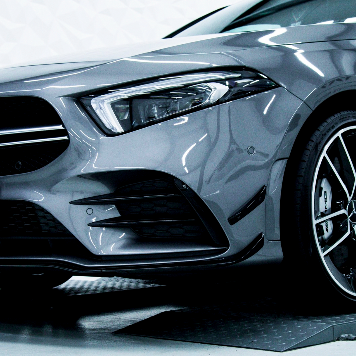 Silver car front with sleek headlight design and a prominent wheel, parked indoors on a metallic floor, reflecting overhead lights.