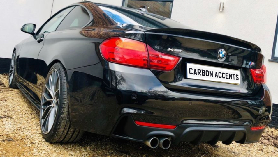 A black sports car with dual exhaust is parked on a gravel driveway beside a white building. The license plate reads "CARBON ACCENTS."