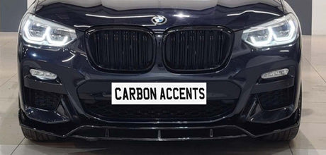 Car front grille with illuminated headlights; BMW logo above, and "CARBON ACCENTS" text on a simulated plate. The car is in a showroom setting with a tiled floor.