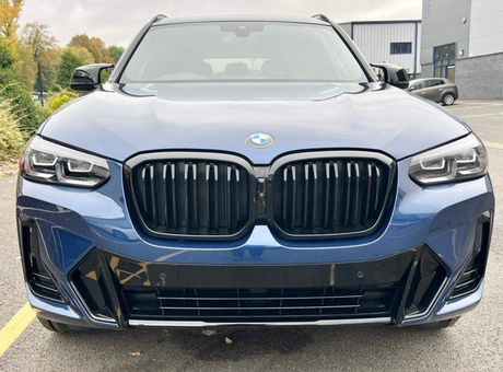 A blue BMW SUV is parked in a parking lot, displaying a prominent front grille and headlights, with trees and industrial buildings in the background.