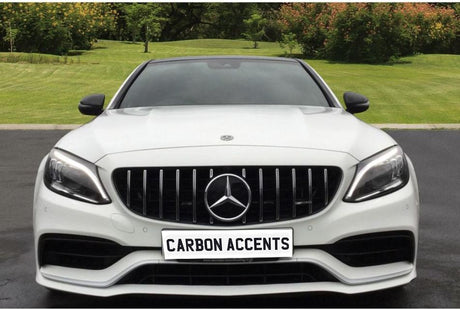 A white car is parked on a driveway, displaying the text "CARBON ACCENTS" on its front plate. The background features a garden with grass and trees.