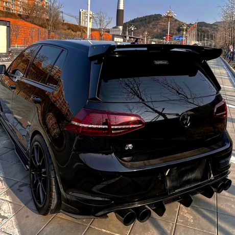 A black car, parked on a tiled pavement, features a prominent rear spoiler and quad exhaust pipes. It's set against an urban backdrop with buildings and hills in the distance.