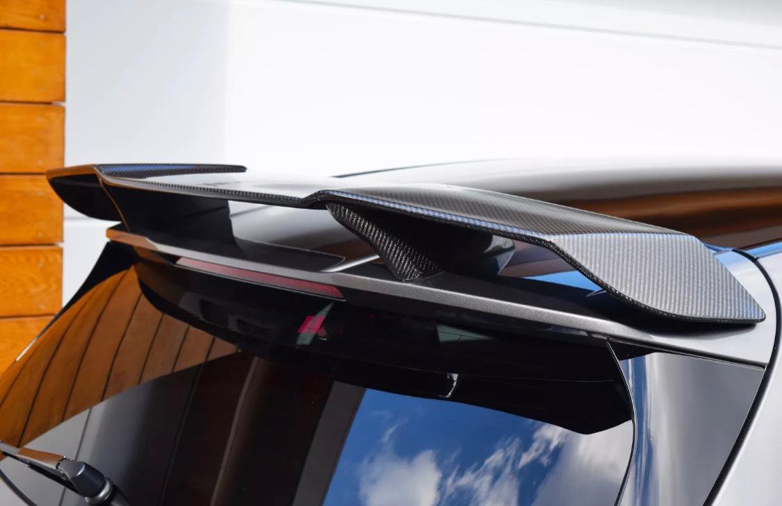 A carbon fiber car spoiler is mounted on the roof of a black vehicle, parked against a wooden structure and white wall, with blue sky reflections visible on the window.