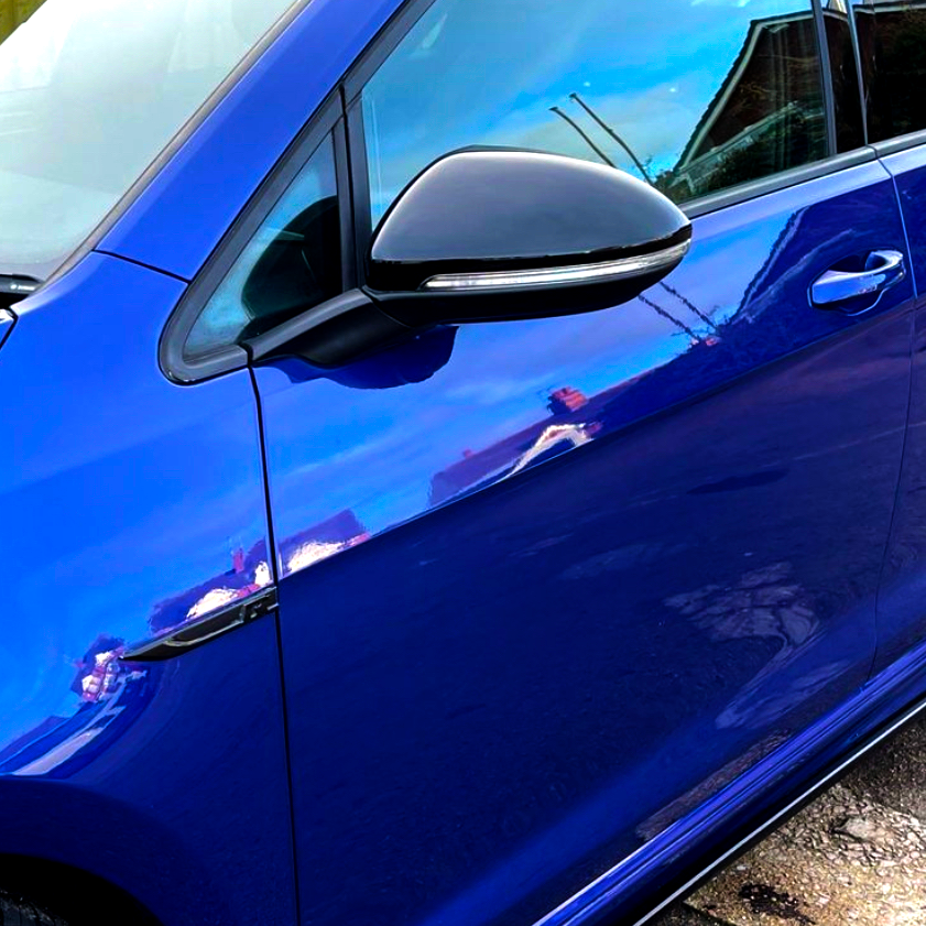 A blue car door with a black side mirror reflects the surrounding houses and sky in a driveway setting, showcasing bright outdoor lighting.