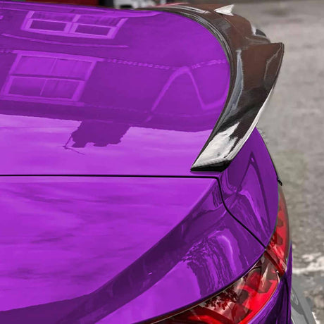 A vibrant purple car with a carbon fiber spoiler is parked, reflecting building windows on its glossy surface. Tail lights are partially visible, hinting at a rear view.