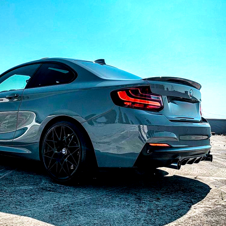 A sleek, gray sports car parked on a concrete surface, showing rear taillights and dark alloy wheels, under a clear blue sky. A warehouse is visible in the background.