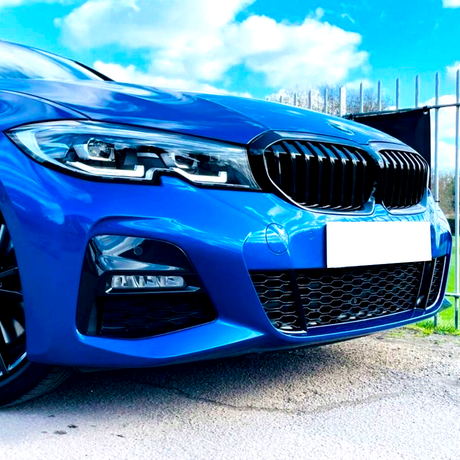 A blue car with a prominent grille and headlights is parked on a paved surface, next to a metal fence under a bright blue sky with scattered clouds.