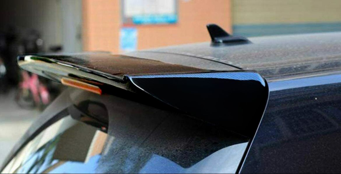 A black car roof spoiler with a reflective finish is mounted on a vehicle, parked in an urban setting. A building's blurred exterior is visible in the background.
