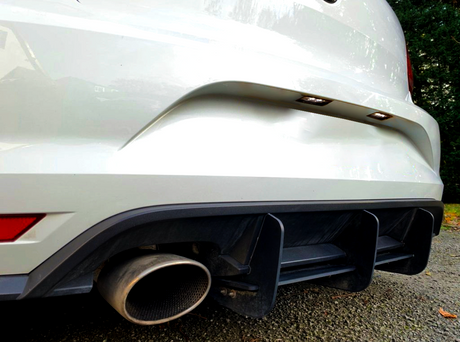 A white car's rear with dual exhaust pipes and a black diffuser sits on a gravel driveway, bordered by greenery, under a clear sky.