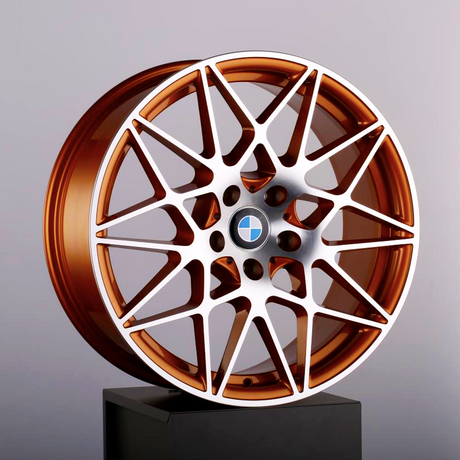 The bronze alloy wheel features an intricate, multi-spoke design, resting on a black pedestal. A blue-and-white circular emblem is at the center, set against a plain background.