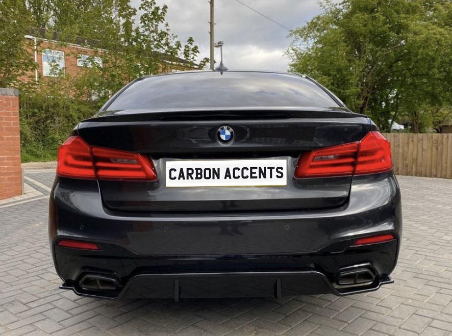 A black BMW car is parked with its back facing the viewer, displaying a license plate reading "CARBON ACCENTS," in a driveway with trees and a brick building nearby.