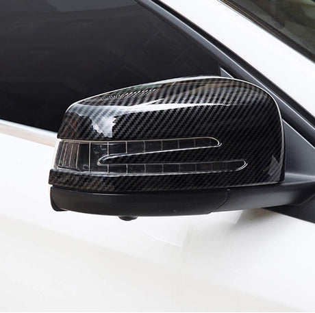A carbon fiber side mirror cover on a white car, displaying a sleek design with integrated indicator lights, positioned against a dark window background.