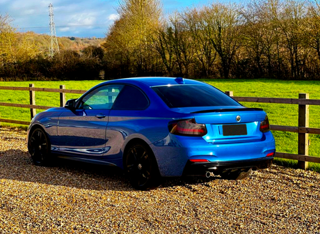 A blue sports coupe is parked on a gravel path, surrounded by a wooden fence and green field, with trees in the background under a partly cloudy sky.