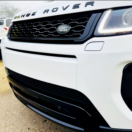 A white Range Rover front grille displays the "LAND ROVER" logo, with sleek headlights and a polished bumper, parked on a paved surface.