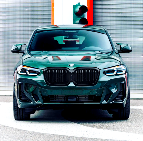 A green BMW car is parked on a circular pavement, positioned in front of a brushed metal wall and a traffic light.