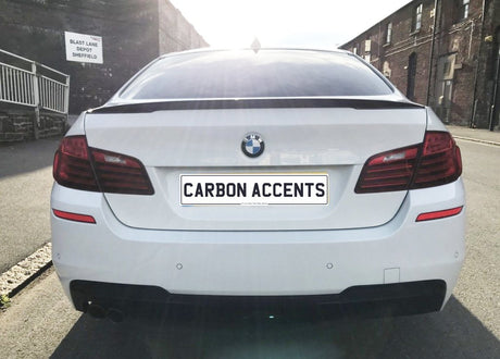 A white BMW sedan is parked on a street, featuring a license plate reading "CARBON ACCENTS." In the background, there's a building with a sign saying "BLAST LANE DEPOT SHEFFIELD."