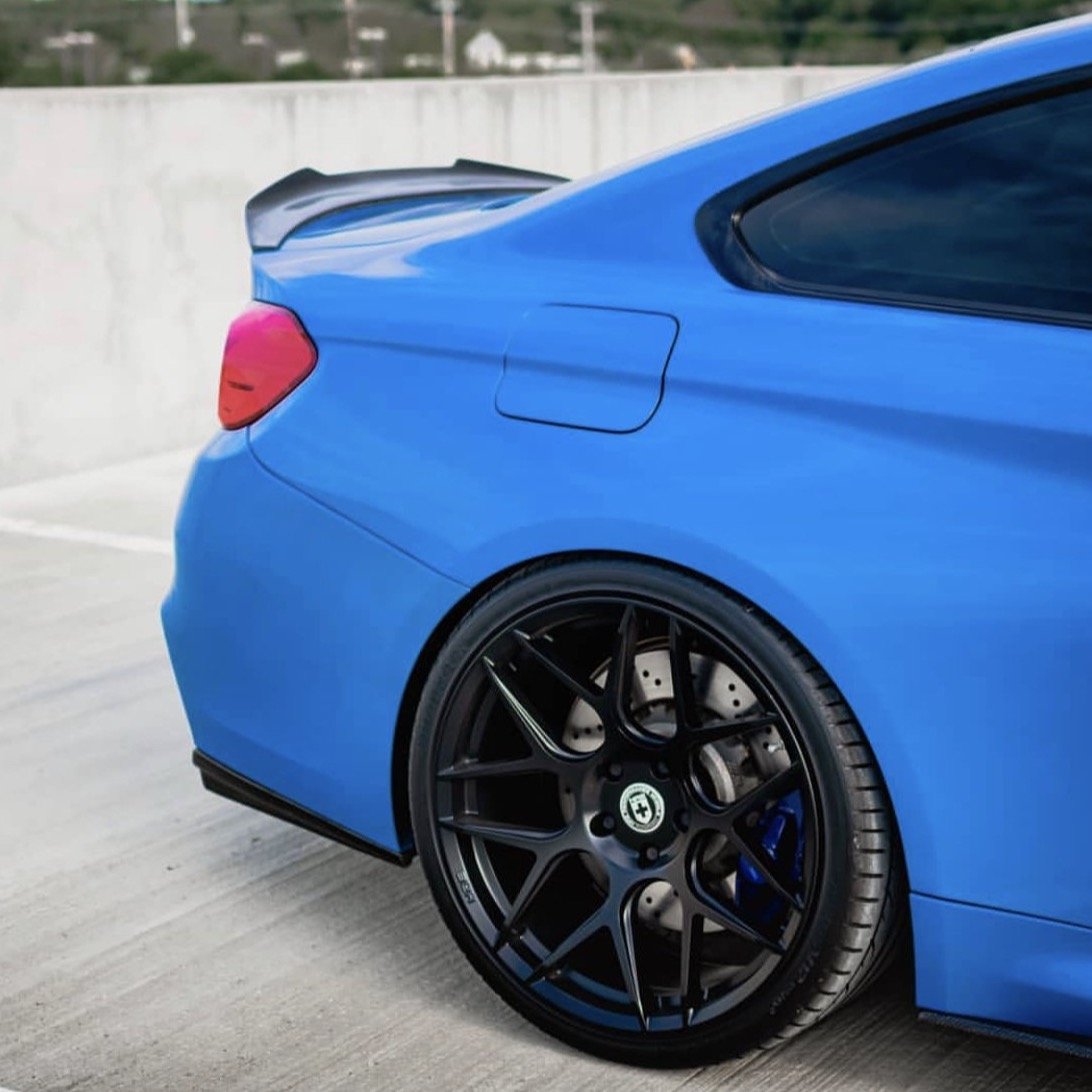 A blue sports car is stationary on a rooftop parking lot, highlighting its rear wheel with glossy black rims and a sleek spoiler.