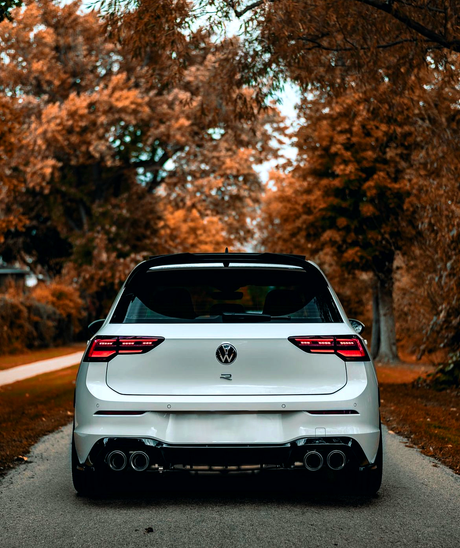 A white car is parked on a narrow road, surrounded by trees with autumn foliage. The car's logo and dual exhaust are visible.