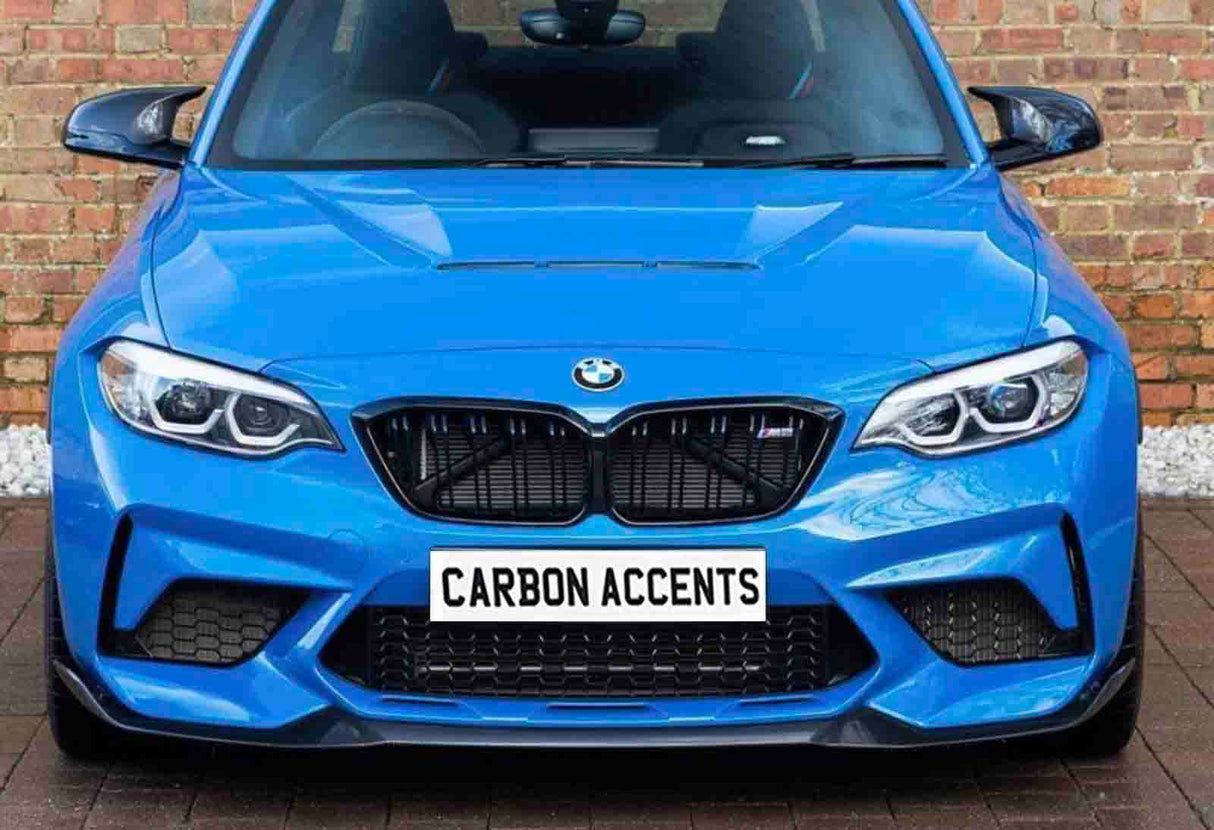 A bright blue BMW car is parked facing forward, displaying a bold front grille and headlights. The license plate reads "CARBON ACCENTS." It is set against a brick wall backdrop.
