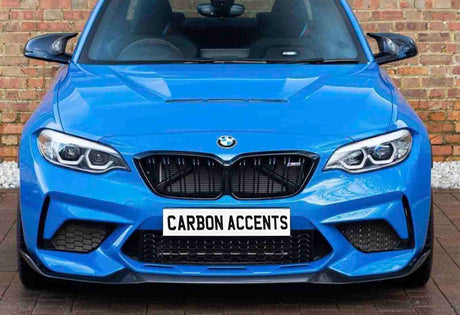 A bright blue BMW car is parked facing forward, displaying a bold front grille and headlights. The license plate reads "CARBON ACCENTS." It is set against a brick wall backdrop.