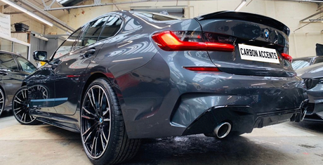 A dark gray sedan with sleek, reflective surfaces is parked indoors. Its license plate reads "CARBON ACCENTS." Other vehicles and industrial lighting are visible in the background.