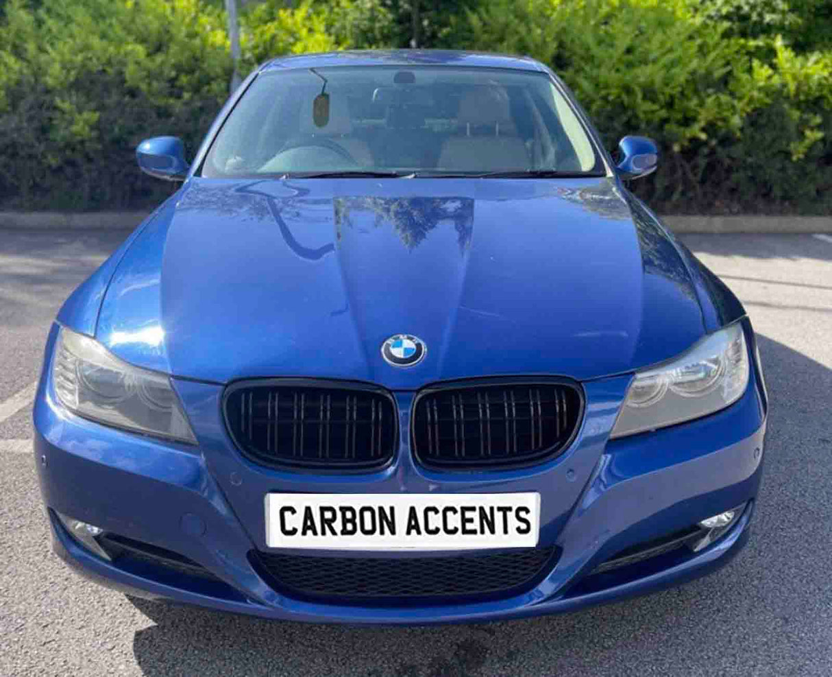 A blue BMW car is parked on a sunny day, facing forward. The license plate reads "CARBON ACCENTS," surrounded by greenery in the background.