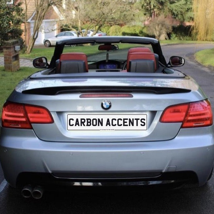 A silver convertible car with a soft top down is parked on a residential street. The license plate reads "CARBON ACCENTS." Trees and houses are visible in the background.