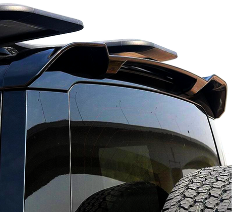A black SUV with a rooftop spoiler and a rear tire mounted on the back, parked outdoors on a sunny day, with a partial view of a curved overpass in the background.