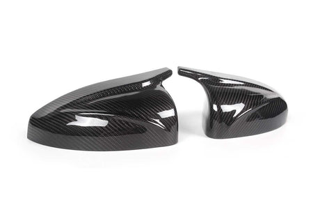 Two sleek, black carbon fiber car mirror covers rest on a plain white surface, reflecting light and showcasing their glossy, patterned finish.