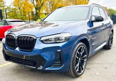 A blue SUV is parked on a paved surface, featuring a prominent black grille and sleek design. Autumn trees with golden leaves form the backdrop, alongside another parked vehicle.