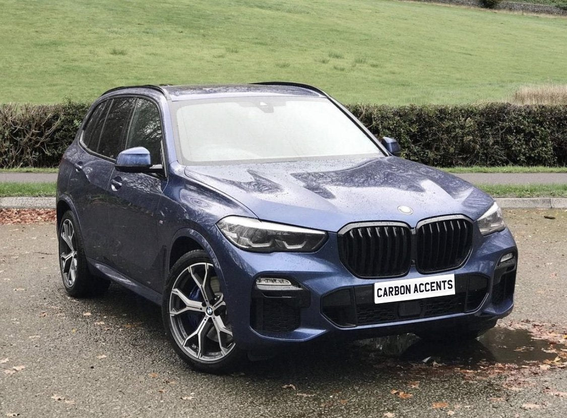 A blue SUV with the text "CARBON ACCENTS" on the front license plate is parked on a wet road, surrounded by grass and bushes.