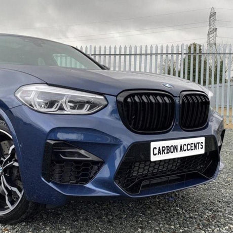 A blue car with a bold front grille and the text "CARBON ACCENTS" on the plate, parked on gravel, with a tall metal fence and overcast sky in the background.