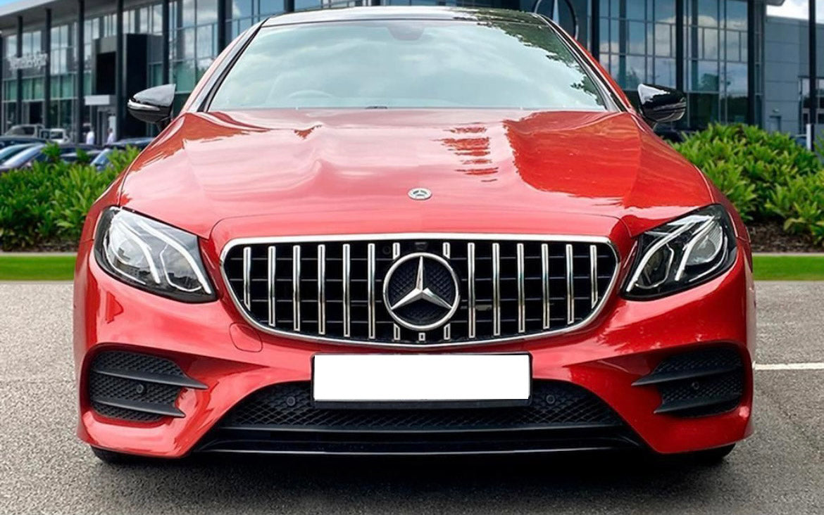 A red car with a distinctive grille and emblem is parked outdoors. Office buildings are visible in the background, along with greenery on either side.